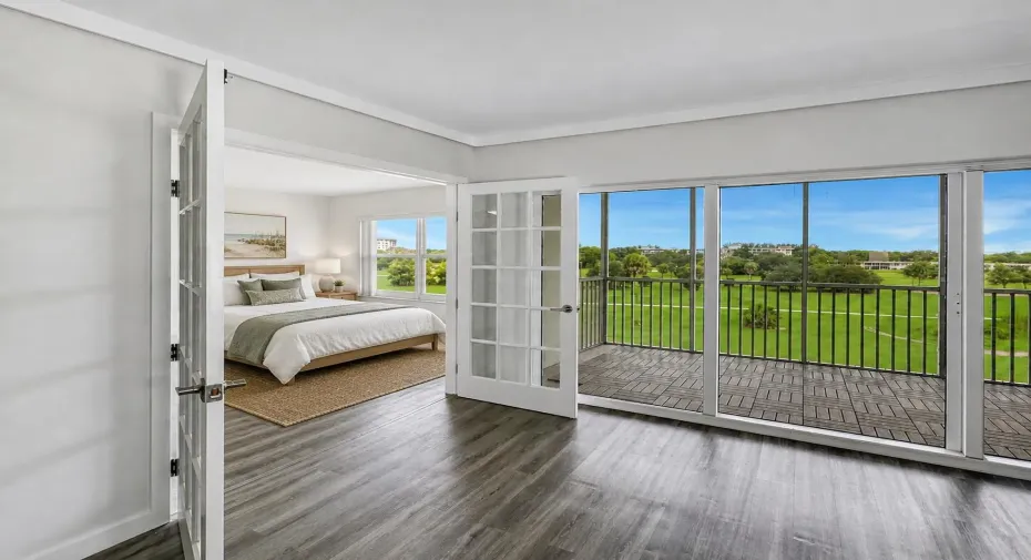 Second Bedroom features French doors opening directly to the living area, creating flexible use for guests, a home office, or additional living space. Vinyl flooring and large windows provide a bright, airy feel with scenic green space views nearby. Photo has been virtually staged for illustrative purposes. Furniture and decor shown are digitally added.