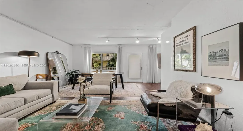 A bright, eclectic living space with white walls and track lighting, featuring a mix of vintage and modern furnishings — a light grey sofa, a dark leather chair, a transparent acrylic coffee table, and layered floral rugs. An Andy Warhol Museum poster and a palm tree photograph add artistic character to the walls, while a large leaning mirror and floor lamp bring depth to the room. A dining area with a dark table is visible toward the back, with natural light streaming through sheer curtains.