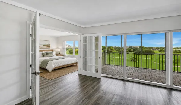 Second Bedroom features French doors opening directly to the living area, creating flexible use for guests, a home office, or additional living space. Vinyl flooring and large windows provide a bright, airy feel with scenic green space views nearby. Photo has been virtually staged for illustrative purposes. Furniture and decor shown are digitally added.