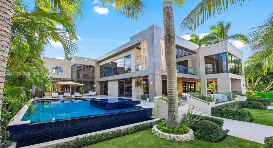 Resort-style pool and spa with dark mosaic tile, sun deck with chaise lounges, pergola, manicured lawn, tropical landscaping, and full two-story modern rear elevation with stone cladding, floor-to-ceiling glass, second-level balcony, and covered loggia.
