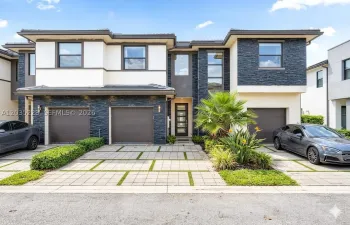 Modern townhome exterior — sleek charcoal stone and white stucco facade, single-car garage, glass-panel entry door, lush tropical landscaping with palms framing a geometric paver driveway.