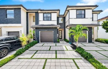 Modern townhome exterior — sleek charcoal stone and white stucco facade, one-car garage, glass-panel entry door, highlighted by landscaping with palms framing a geometric paver driveway.