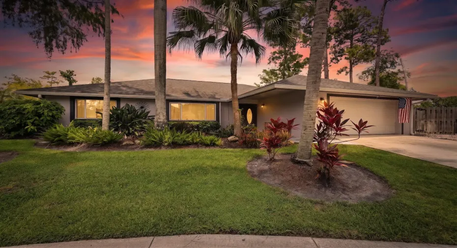 Front exterior of a single-story South Florida home with lush tropical landscaping, palm trees, two-car garage, wide driveway, warm interior lighting, and beautiful sunset curb appeal.
