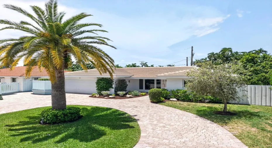 Exterior residence featuring a paver driveway, mature palm tree, lush landscaping, white stucco facade, and a tile roof