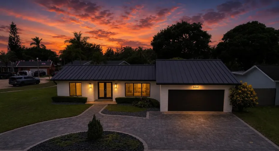 Contemporary exterior featuring a dark standing seam metal roof, white painted brick facade, a two-car garage, and a paver driveway