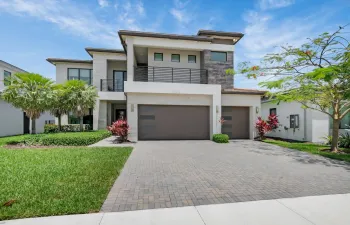 Contemporary two-story residence featuring a paver driveway, multiple garage doors, a second-story balcony with metal railing, and a stone accent facade