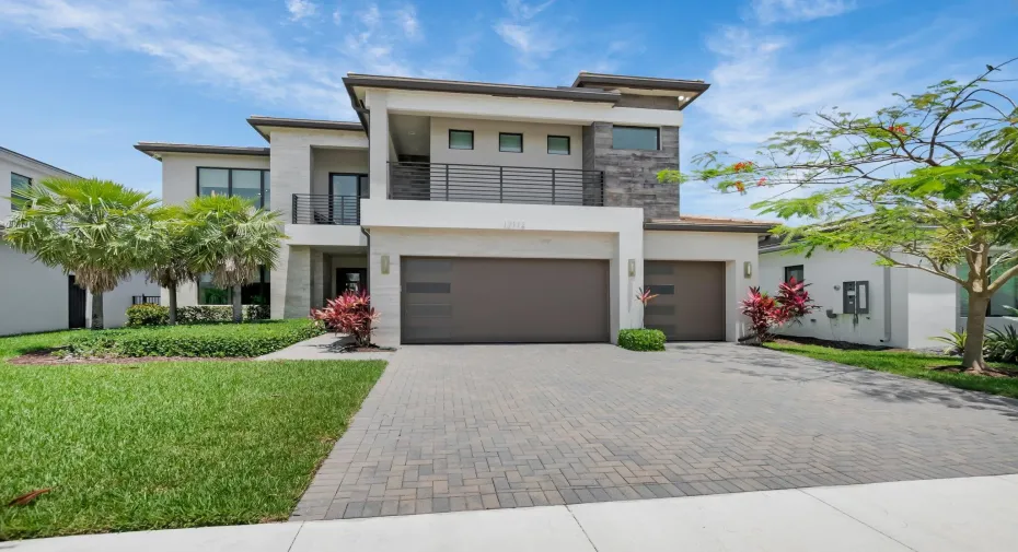 Contemporary two-story residence featuring a paver driveway, multiple garage doors, a second-story balcony with metal railing, and a stone accent facade