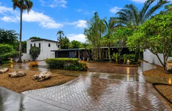 Paved driveway featuring a herringbone pattern, gravel landscaping with natural rock elements, mature palm trees, and lush tropical foliage