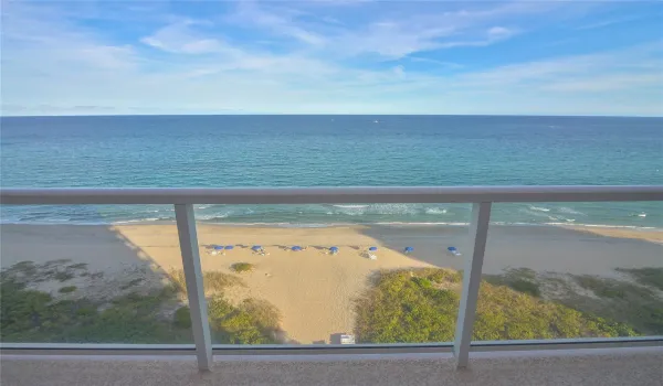 Oceanfront balcony featuring a glass railing, expansive ocean, and sandy beach with natural vegetation
