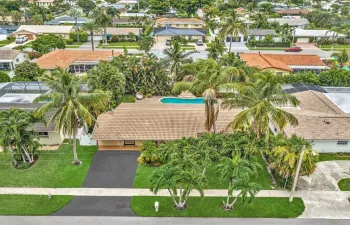 Expansive green lawn, mature palm trees, asphalt driveway, exterior light brown tile roof, and a visible swimming pool