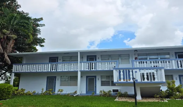 Two-story building featuring white exterior, blue trim accents, multiple balconies with white and blue balusters, and a green lawn with landscaping