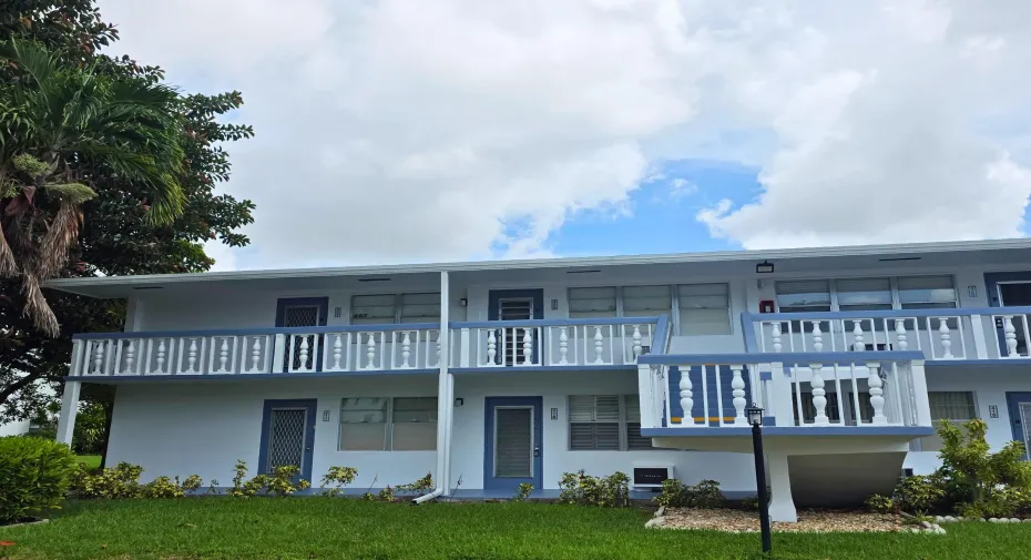Two-story building featuring white exterior, blue trim accents, multiple balconies with white and blue balusters, and a green lawn with landscaping