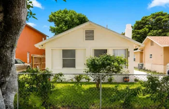 Single-story exterior with a light-colored facade, fenced front yard, and mature greenery