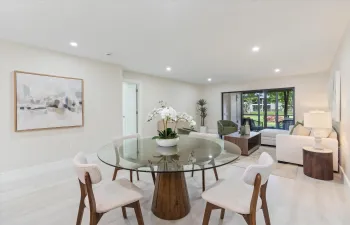Modern open-concept dining and living area featuring a round glass-top table with wood-finish pedestal base, paired upholstered dining chairs, recessed ceiling lighting, light wood-finish flooring, and expansive glass doors opening to landscaped outdoor spaces with mature greenery