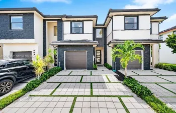 Modern townhome exterior — sleek charcoal stone and white stucco facade, one-car garage, glass-panel entry door, highlighted by landscaping with palms framing a geometric paver driveway.