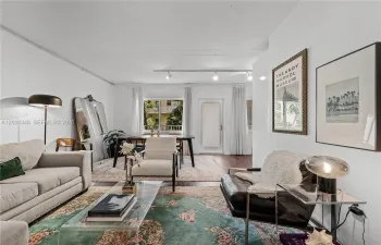 A bright, eclectic living space with white walls and track lighting, featuring a mix of vintage and modern furnishings — a light grey sofa, a dark leather chair, a transparent acrylic coffee table, and layered floral rugs. An Andy Warhol Museum poster and a palm tree photograph add artistic character to the walls, while a large leaning mirror and floor lamp bring depth to the room. A dining area with a dark table is visible toward the back, with natural light streaming through sheer curtains.