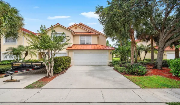 Two-story residence with a metal roof accent, featuring a two car garage, and driveway, and professionally landscaped grounds with mature palms, flowering shrubs, and established trees throughout the property