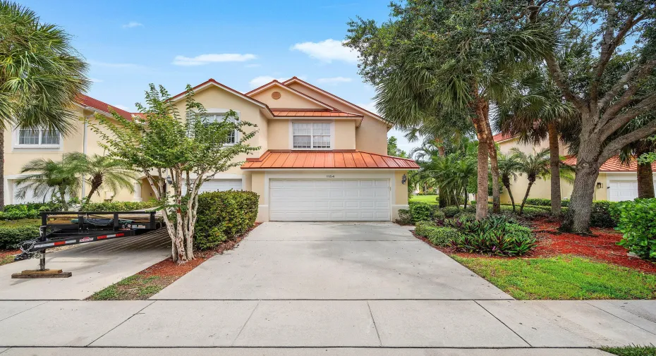 Two-story residence with a metal roof accent, featuring a two car garage, and driveway, and professionally landscaped grounds with mature palms, flowering shrubs, and established trees throughout the property
