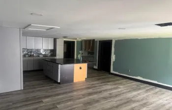 Modern open-concept space featuring a wood-finish plank floor throughout, contemporary kitchen with dark cabinetry and a central island with warm-toned accent paneling, recessed ceiling lighting, and a large chalkboard wall ideal for creative or organizational use