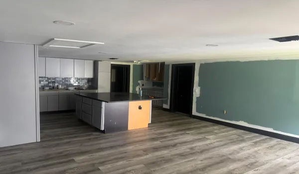 Modern open-concept space featuring a wood-finish plank floor throughout, contemporary kitchen with dark cabinetry and a central island with warm-toned accent paneling, recessed ceiling lighting, and a large chalkboard wall ideal for creative or organizational use