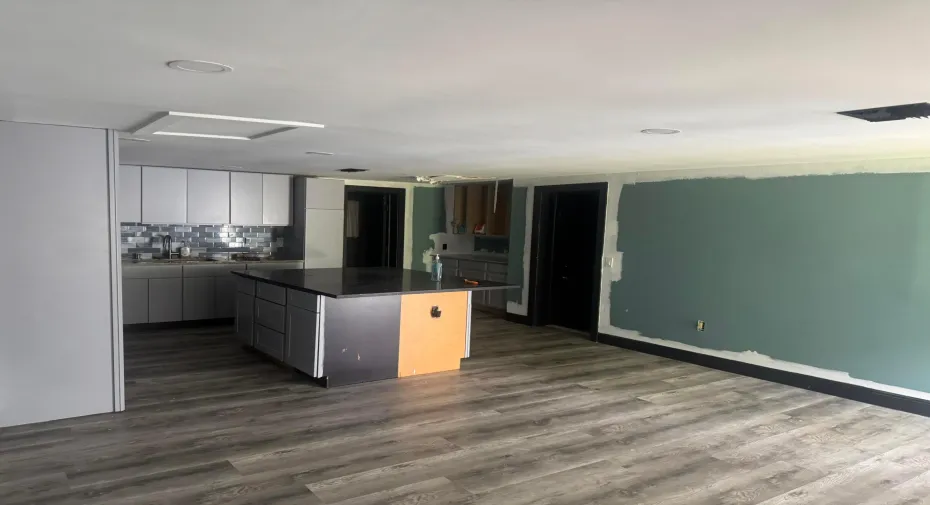 Modern open-concept space featuring a wood-finish plank floor throughout, contemporary kitchen with dark cabinetry and a central island with warm-toned accent paneling, recessed ceiling lighting, and a large chalkboard wall ideal for creative or organizational use