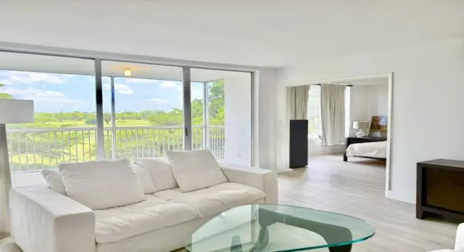 Bright, open-concept living area showcasing floor-to-ceiling sliding glass doors, light wood-finish flooring, and expansive panoramic views of surrounding landscaping