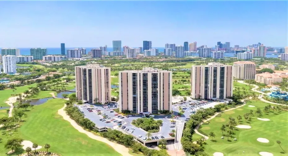 Three high-rise buildings situated amidst a golf course landscape with a distant city skyline and ocean background