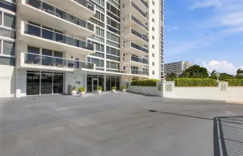 High-rise exterior featuring concrete structural design, glass-paneled balconies, floor-to-ceiling glass entry doors, and a paved arrival court