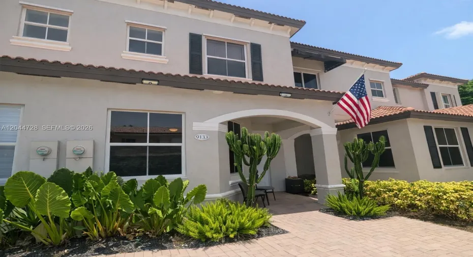Charming two-story residence with a clean exterior and well-maintained details. Features a tiled roof, white-framed windows, and an elegant arched entrance leading to a tropical garden with tall succulents and ornamental plants. Located in a quiet and attractive condominium.
