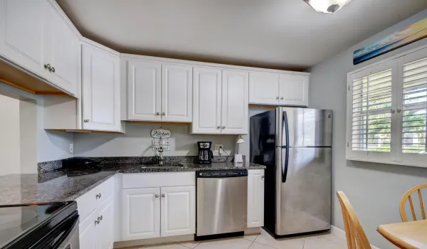 White cabinetry, dark stone countertops, stainless steel appliances, tile flooring, and a window with plantation shutters