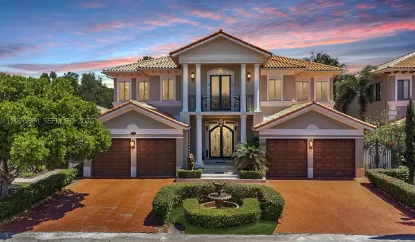 A grand Mediterranean-style estate at sunset, with a terracotta tile roof, ornate double entry doors, lion statues, Corinthian columns, a Juliet balcony, and a circular fountain driveway flanked by manicured hedges. Stately and impressive.