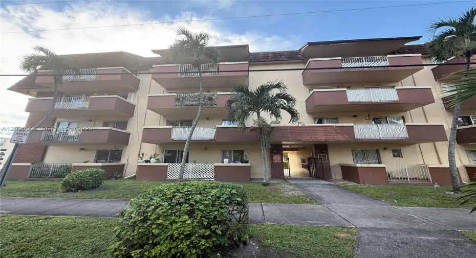 Multi-story exterior featuring individual private balconies with white railings, concrete walkways, and mature palm trees
