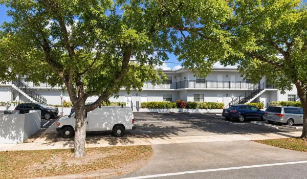 Two-story multi-unit structure featuring light-toned exterior finish, second-level balcony with metal railing, designated surface parking, and mature trees