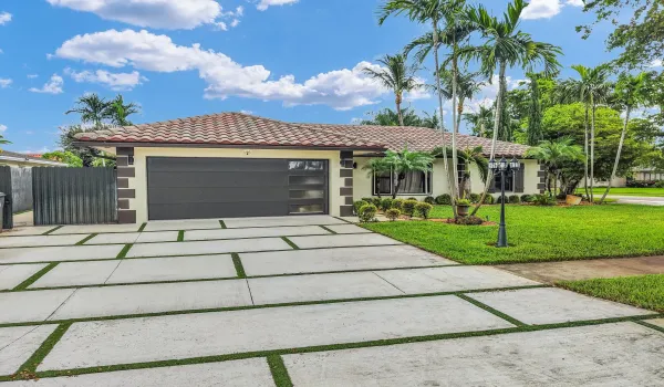 Terracotta-style roof tiles, stucco exterior, modern garage door with frosted panels, patterned concrete driveway with artificial turf accents, and established landscaping with palm trees