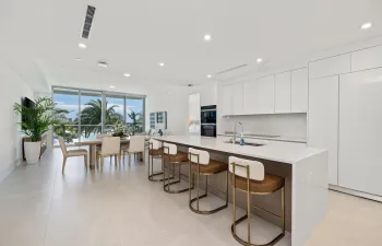 Sleek kitchen with white cabinetry and integrated appliances, expansive central island with countertop seating, open-concept living space, extensive floor-to-ceiling windows, and recessed lighting