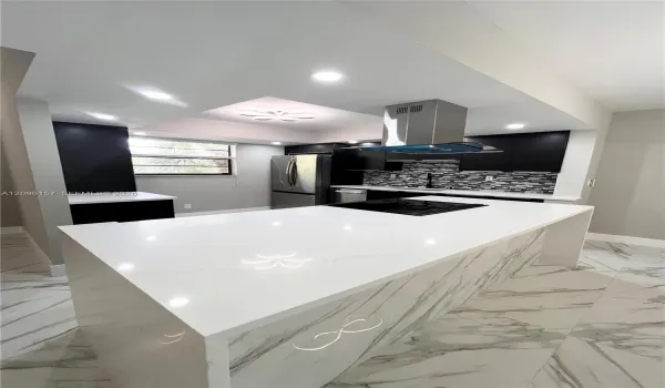 Kitchen featuring a central island with a waterfall-edge countertop, black cabinetry, stainless steel range hood, stone-patterned tile flooring, and a mosaic backsplash