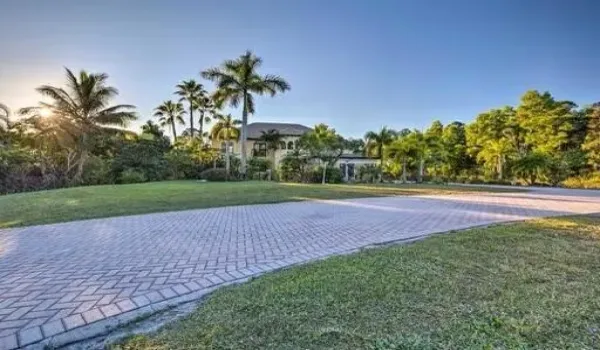 Expansive paver driveway leading to a multi-story residence with a stucco exterior, surrounded by mature palm trees and lush greenery