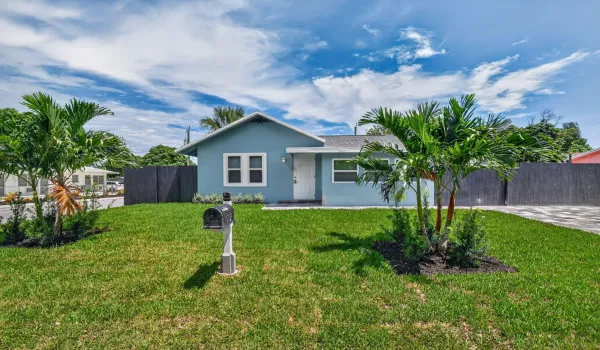 Light blue exterior with white trim, well-maintained lawn, mature palm trees, and a dark privacy fence