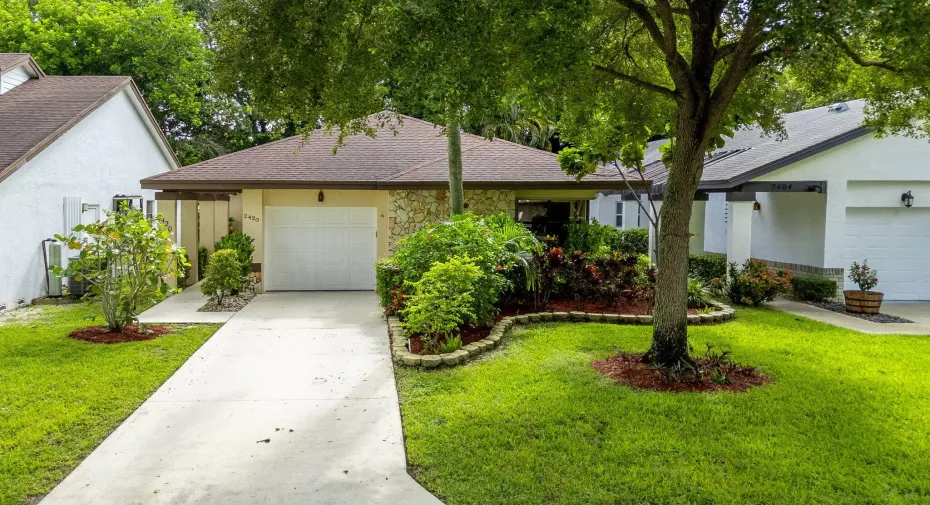 Single-story residence featuring a paved driveway, attached garage, stone veneer facade, mature trees, and landscaped grounds
