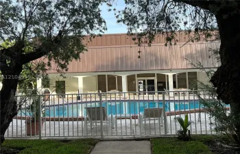 Swimming pool with brick coping, surrounded by a paved deck and enclosed by a metal perimeter fence, featuring a covered patio with structural columns