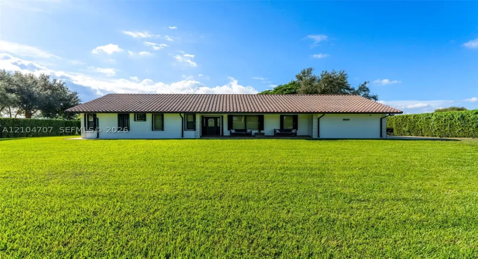 Single-story residence featuring a low-pitched tiled roof, light-toned exterior finish, and an expansive manicured lawn bordered by mature hedge fencing