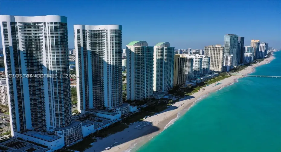 Coastal high-rise residences with private balconies, set along a sandy beach and turquoise waters