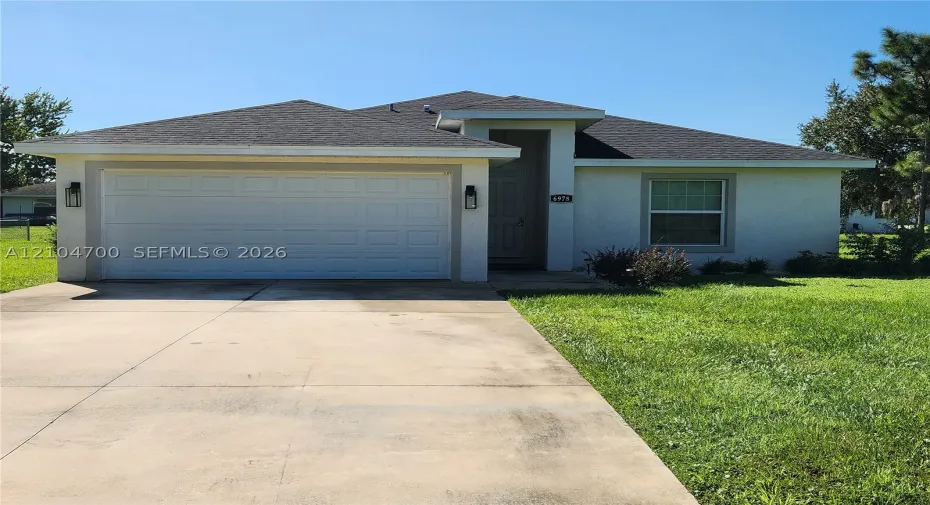 Stucco exterior with a shingled roof, featuring an attached garage, concrete driveway, and a front entrance with a porch light
