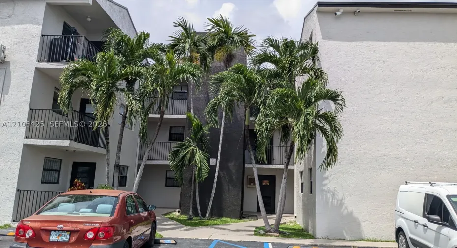 Multi-story building featuring balconies with black metal railings, textured light-colored stucco exterior, and mature palm trees