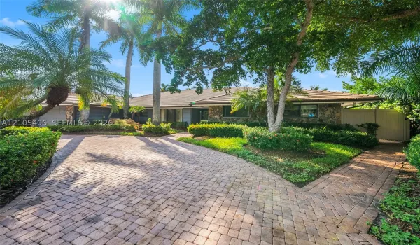 Stone-clad exterior with a tiled roof, paver driveway, and lush landscaping, featuring mature palm trees and established hedging