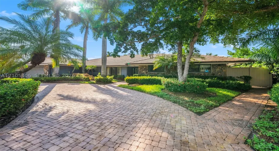 Stone-clad exterior with a tiled roof, paver driveway, and lush landscaping, featuring mature palm trees and established hedging