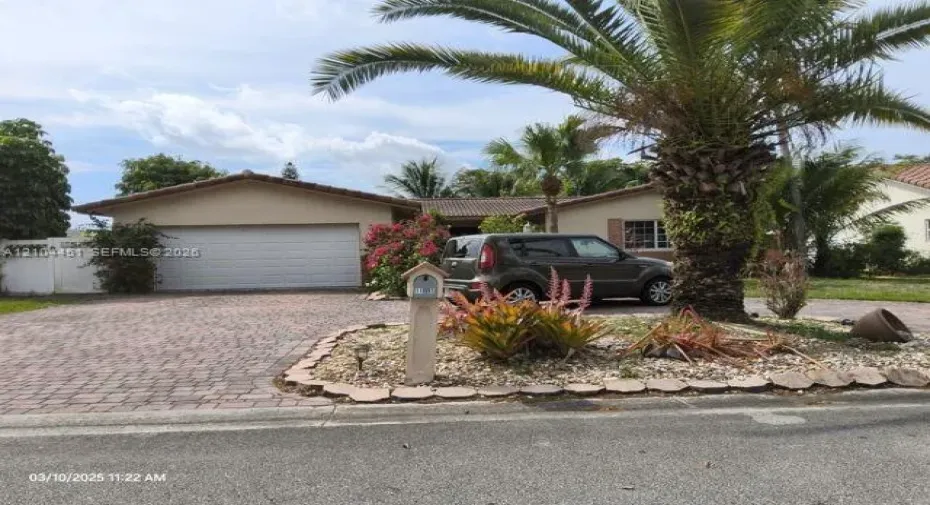 Paved brick driveway, attached two-car garage, mature palm trees, and established landscaping