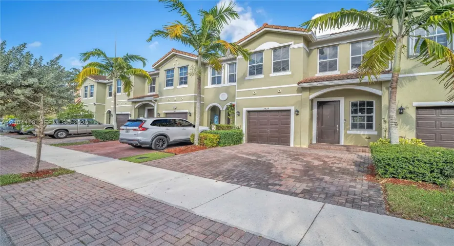 Stucco exterior with arched entryway, paver driveway, brown garage doors, and mature palm trees