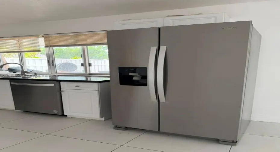 Stainless steel side-by-side refrigerator, stainless steel dishwasher, white cabinetry, dark stone countertop, and tile flooring