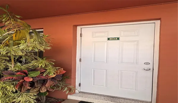 White paneled entry door with chrome hardware, set against a terracotta-toned exterior with integrated planter featuring vibrant foliage